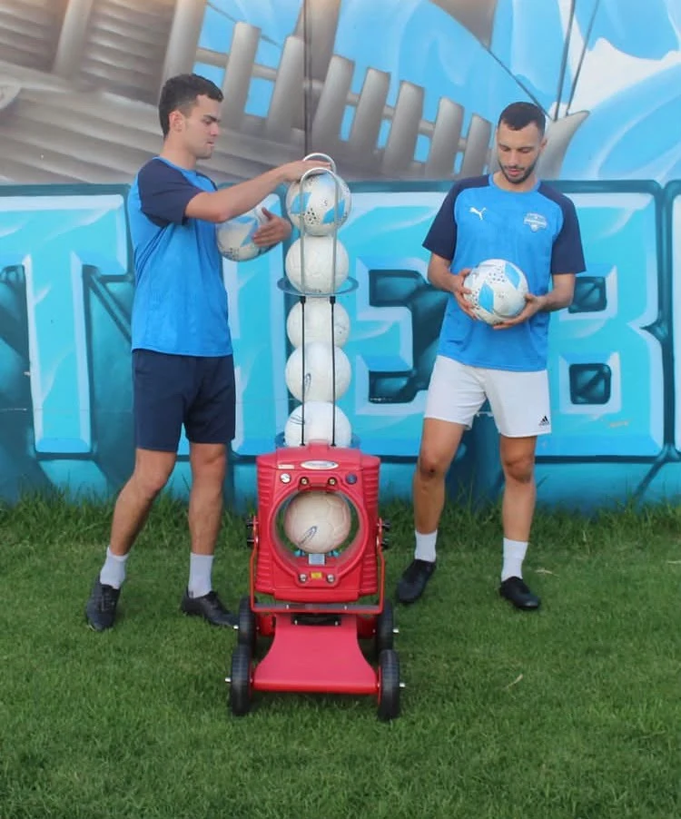 Two players beside a red POWAPASS machine, with one loading footballs into the chute